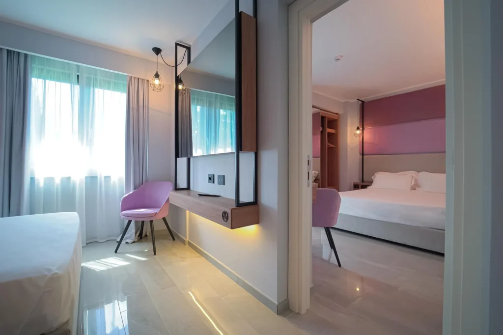 Nell'immagine si vede una camera con letto matrimoniale. Le pareti sono bianche e rosa. Di fronte al letto un tavolino rotondo in legno e due poltroncine rosa in velluto. Si intravede anche la seconda stanza.